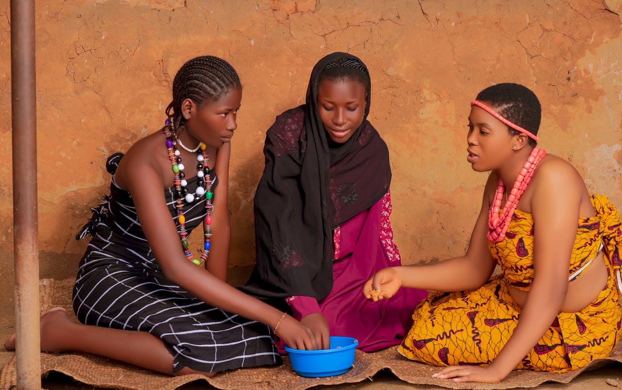Three African women in traditional attire sit together, engaged in conversation.
